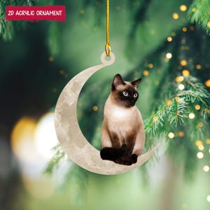 Könnte beinhalten: Ein Weihnachtsbaumschmuck mit einer Siamkatze, die auf einem Halbmond sitzt. Die Katze hat einen beigefarbenen Körper, dunkelbraune Punkte und blaue Augen. Der Mond ist hellbeige, und der Schmuck hängt an einer goldenen Schnur. Der Text "2D ACRYLIC ORNAMENT" befindet sich in einem roten Feld.