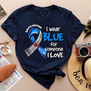 Puede incluir: Camiseta azul marino con el texto "I wear BLUE for someone I LOVE" y una cinta azul con una gota roja, promoviendo la concienciación sobre la diabetes. Las palabras "Diabetes awareness" también están impresas en la camiseta.
