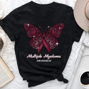 May include: Black t-shirt featuring a burgundy butterfly design composed of smaller butterflies, with a burgundy ribbon in the center. The words "Multiple Myeloma Awareness" are printed in white.