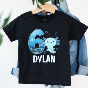 Könnte beinhalten: Schwarzes T-Shirt mit einer blauen Zahl 6, einem Axolotl und dem Namen "DYLAN" darauf gedruckt.