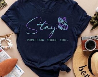 May include: Navy blue t-shirt with the word "Stay" in a gradient of blue and purple, along with a butterfly graphic. The text "Tomorrow Needs You" is below the word "Stay".
