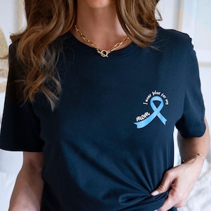 Puede incluir: Camiseta azul marino con un lazo azul claro y el texto "I wear blue for my mom". La camiseta tiene cuello redondo y mangas cortas. El diseño está en el pecho izquierdo.