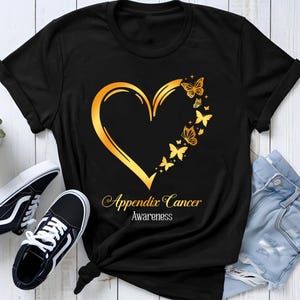 May include: A black short-sleeved t-shirt featuring a gold graphic of a heart outline formed by butterflies on the right side. Below the heart, the text reads Appendix Cancer Awareness in a gold script and white sans-serif font.