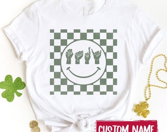 Puede incluir: Una camiseta blanca con un gráfico verde salvia de una cara sonriente con manos formando las letras "BABY" en lenguaje de señas sobre un fondo a cuadros. Un recuadro rojo con el texto blanco "CUSTOM NAME" aparece en la parte inferior.