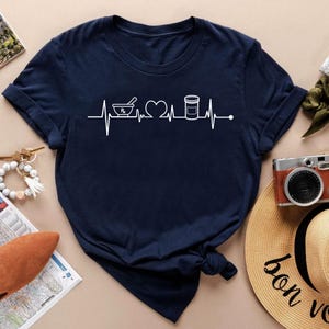 Könnte beinhalten: Ein marineblaues T-Shirt mit einer weißen Grafik eines Herzschlags mit einem Mörser und Stößel, einem Herzen und einer Pillenflasche.