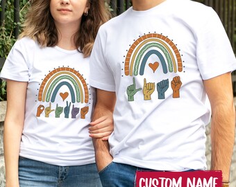 Puede incluir: Dos camisetas blancas con un gráfico de un arcoíris multicolor con un corazón y manos formando letras en lenguaje de señas. El diseño incluye rayas en tonos tierra y formas de manos. Un recuadro rojo muestra el texto CUSTOM NAME.