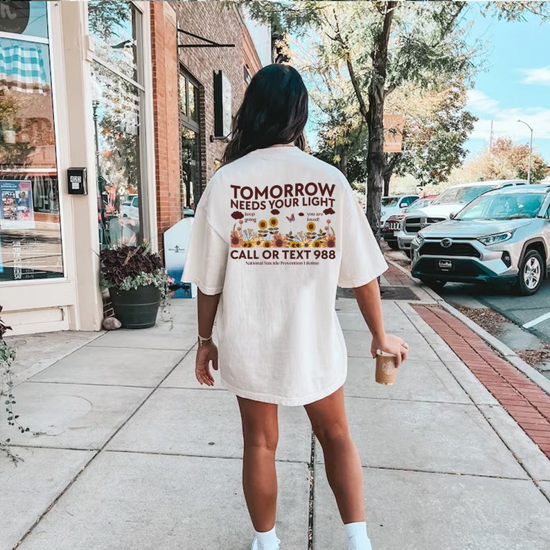 Pu&ograve; includere: Maglietta bianca oversize con una stampa grafica di girasoli, farfalle e nuvole. Il testo sulla maglietta recita "Tomorrow Needs Your Light" e "Call or Text 988 - National Suicide Prevention Lifeline".