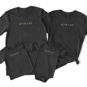 May include: Four dark grey t-shirts and a onesie with the text "AIR FORCE DAD", "AIR FORCE MOM", "AIR FORCE BRO", and "AIR FORCE SIS". The shirts have a crew neck design and are made of a soft material.