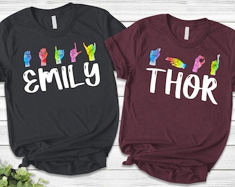 May include: A black t-shirt with the name EMILY and a maroon t-shirt with the name THOR printed in white. Above each name are colourful, watercolour-style hand signs representing the letters of the names in American Sign Language.