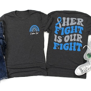 Puede incluir: Dos camisetas de color gris oscuro. Una presenta un diseño de arcoíris y cinta azul con el texto "Custom Text". La otra camiseta tiene el texto "HER FIGHT IS OUR FIGHT" en azul y gris, con un gráfico de cinta azul.