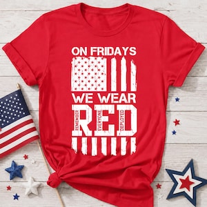 Könnte beinhalten: Ein leuchtend rotes Kurzarm-T-Shirt mit weißem, abgenutztem Grafiktext, der ON FRIDAYS WE WEAR RED REMEMBER EVERYONE DEPLOYED lautet, begleitet von einer stilisierten weißen Grafik der amerikanischen Flagge mit Sternen und Streifen.