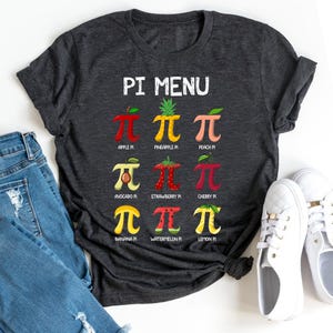 Op de afbeelding: Een donkergrijs T-shirt met een witte afbeelding van een fruit-thema "Pi Menu". De afbeelding toont een reeks verschillende vruchten, elk met een "pi"-symbool, en de naam van de fruittaart eronder.