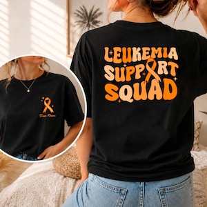 Könnte beinhalten: Ein schwarzes T-Shirt mit einer orangefarbenen Schleife auf der linken Brust und dem Text Team Name. Auf der Rückseite steht LEUKEMIA SUPPORT SQUAD in großen, orangefarbenen Retro-Buchstaben, ergänzt durch eine orange Schleife und Sterne.