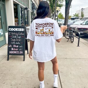 Pu&ograve; includere: Maglietta bianca con una grafica di girasoli e farfalle. Il testo "Domani ha bisogno della tua luce" e "Chiama o invia un messaggio al 988" &egrave; stampato sulla maglietta.