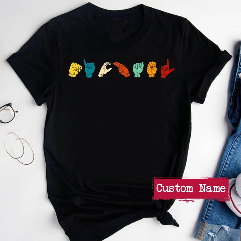 May include: Black t-shirt with a row of colorful hand signs in American Sign Language spelling "I love you". The text "Custom Name" is printed on the bottom right corner of the shirt.