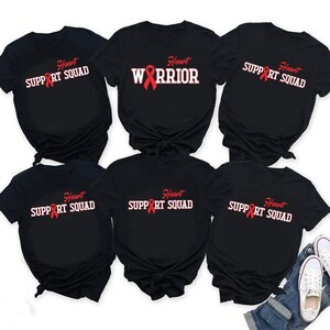 Könnte beinhalten: Schwarze T-Shirts mit weißem und rotem Text. Die Shirts zeigen die Aufschriften "Heart Support Squad" und "Heart Warrior" mit einer roten Schleife. Die Shirts sind kurzärmlig und scheinen aus Baumwolle zu sein.