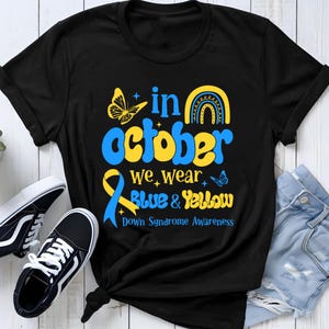 Op de afbeelding: Een zwart t-shirt met een grafisch ontwerp met de tekst "in october we wear blue & yellow" en "Down Syndrome Awareness". Het ontwerp bevat een gele vlinder, een blauwe en gele regenboog en een blauw en geel bewustzijnslintje.