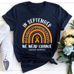 May include: Navy blue t-shirt with the text "IN SEPTEMBER WE WEAR ORANGE" and a rainbow design in orange. The shirt includes a leukemia awareness ribbon. White sneakers and flowers are in the background.