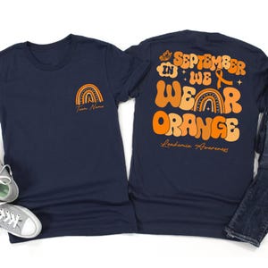 May include: Navy blue t-shirts with orange graphics. One shirt features a rainbow design and the text "Team Name." The other shirt has the text "September in We Wear Orange Leukemia Awareness."