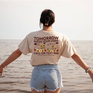 Pu&ograve; includere: Una maglietta beige con il testo "TOMORROW NEEDS YOUR LIGHT" in lettere marroni. La maglietta presenta anche una grafica di girasoli, farfalle e nuvole con il testo "keep going" e "you are loved". La parte inferiore della maglietta recita "CALL OR TEXT 988 National Suicide Prevention Lifeline".