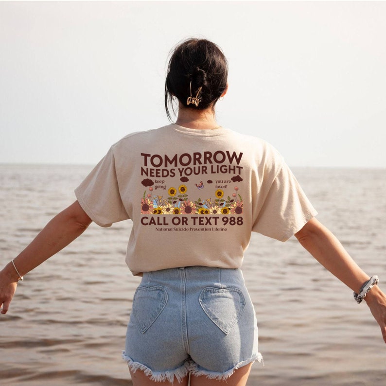 Pu&ograve; includere: Una maglietta beige con il testo "TOMORROW NEEDS YOUR LIGHT" in lettere marroni. La maglietta presenta anche una grafica di girasoli, farfalle e nuvole con il testo "keep going" e "you are loved". La parte inferiore della maglietta recita "CALL OR TEXT 988 National Suicide Prevention Lifeline".
