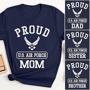 以下が含まれることがあります： ネイビーブルーのTシャツ。白い文字で「PROUD」と書かれ、その下に米空軍のエンブレムがあります。エンブレムの下には「MOM」の文字がプリントされています。追加のデザインには、同じデザインの「DAD」、「SISTER」、「BROTHER」が含まれています。