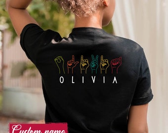 May include: A black t-shirt featuring a colourful graphic on the back. The design displays six hand signs representing the American Sign Language alphabet to spell out the name OLIVIA, with each hand outline in a different vibrant colour.