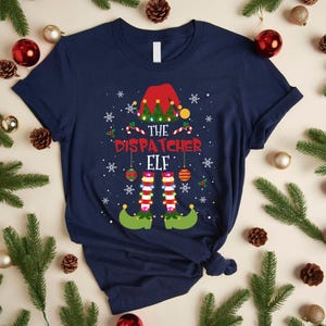 Könnte beinhalten: Marineblaues T-Shirt mit weihnachtlichem Design. Das Design zeigt einen Elfenhut, Zuckerstangen, Schneeflocken und den Text "THE DISPATCHER ELF". Die Beine und Füße des Elfen sind ebenfalls abgebildet.