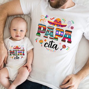 以下が含まれることがあります： カラフルなメキシカン風のデザインが施された白いTシャツで、「パパ・シータ」と書かれています。それに合わせて、カラフルなメキシカン風のデザインが施された白いロンパースで、「ミニ・シータ」と書かれています。