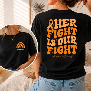 Può includere: Una t-shirt nera con grafiche arancioni. Il retro mostra il testo "HER FIGHT IS OUR FIGHT" con un nastro di sensibilizzazione e la scritta "Leukemia Awareness". Sul petto è presente una piccola icona arcobaleno arancione con "Custom Text".