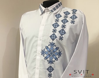 Könnte beinhalten: Ein weißes Langarmhemd mit blauen, gestickten geometrischen Mustern an Kragen, Manschetten und Vorderseite. Die Stickerei zeigt ein kreuzförmiges Design mit einem floralen Motiv. Das Hemd hat einen Knopfleisten vorne und ist mit "SVIT VYSYYVANOK" beschriftet.