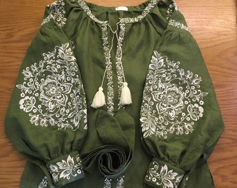 May include: A green linen blouse with white embroidered floral patterns. The blouse has long sleeves with a gathered cuff and a round neckline. The blouse has a tie at the neckline with white tassels.