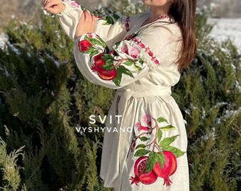 May include: A white linen dress with embroidered pomegranates and green leaves. The dress has a white belt and the text "SVIT VYSVYVANO" is embroidered on the front.