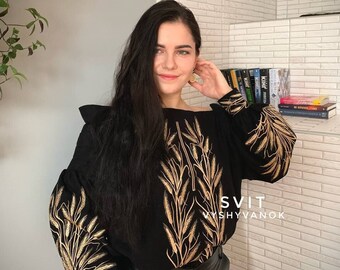 May include: Black long-sleeve shirt with gold embroidery of wheat stalks. The shirt has a slightly off-the-shoulder neckline and a relaxed fit. The text "SVIT VYSYVANOX" is printed on the bottom right corner of the shirt.
