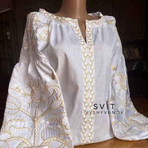 Op de afbeelding: Witte linnen blouse met geborduurde bloemenpatroon in goud- en zilverdraad. De borduurwerk toont een gestileerde boom met bladeren en bloemen. De blouse heeft een V-hals en lange mouwen. De tekst "SVIT VYSVYVANOK" is geborduurd op de voorkant van de blouse.