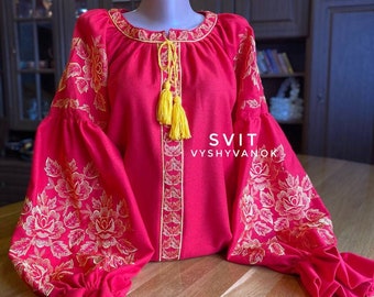 May include: A red embroidered blouse with gold floral designs and yellow tassels. The blouse has long sleeves with ruffles at the cuffs. The text "SVIT VYSYVANOX" is embroidered on the front of the blouse.
