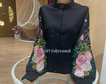 May include: Black button-down shirt with floral embroidery on the sleeves. The embroidery features pink and white roses with green leaves. The shirt has a high collar and long sleeves. The text "SvitVyshyvanok" is visible on the sleeve.