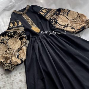 May include: Black dress with gold embroidery on the sleeves and bodice. The dress features a high neckline, long sleeves, and a flowing skirt. The sleeves have a floral pattern. The text "SvitVyshyvanok" is visible on the dress.