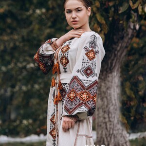 May include: A woman wearing a white embroidered dress with orange and brown geometric patterns. The dress has a belt and long sleeves. The text "SVIT VYSHYVANOK" is visible at the bottom of the image.