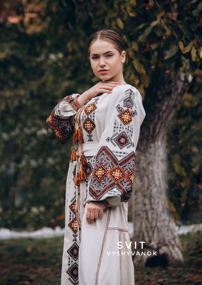 May include: A woman wearing a white embroidered dress with orange and brown geometric patterns. The dress has a belt and long sleeves. The text "SVIT VYSHYVANOK" is visible at the bottom of the image.