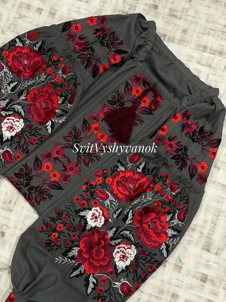 A gray long-sleeved blouse featuring vibrant red, white, and black floral embroidery on the chest and sleeves, with a dark red drawstring tassel at the neckline.