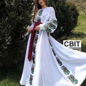 A woman wearing a long white dress with colorful floral embroidery on the chest, sleeves, and skirt, cinched at the waist with a dark red sash.