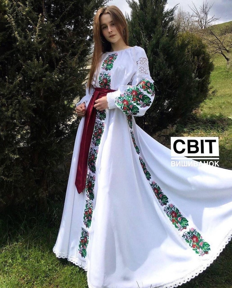 A woman wearing a long white dress with colorful floral embroidery on the chest, sleeves, and skirt, cinched at the waist with a dark red sash.