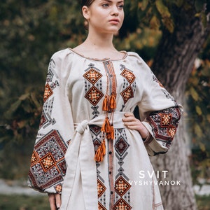 May include: A white linen dress with orange and black embroidery. The dress has a long, flowing skirt and a fitted bodice. The embroidery is in a geometric pattern and features tassels. The dress is labelled "SVIT VYSVYVANOK".