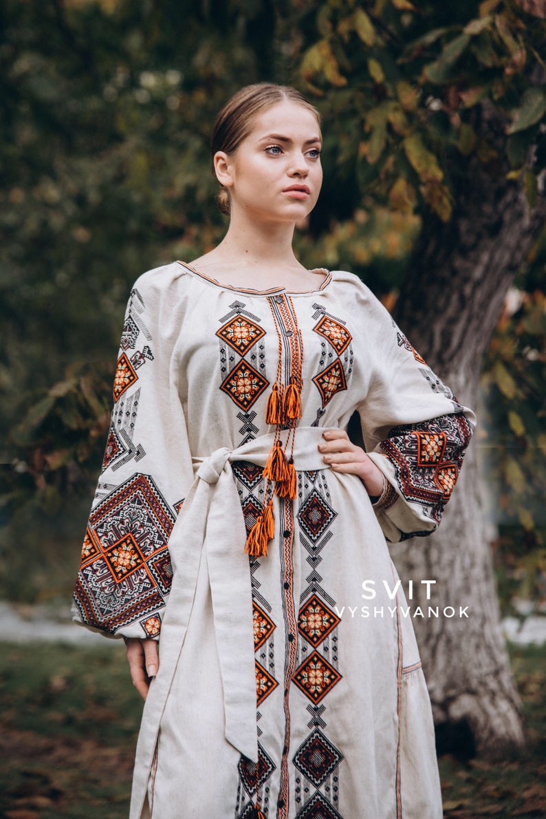 May include: A white linen dress with orange and black embroidery. The dress has a long, flowing skirt and a fitted bodice. The embroidery is in a geometric pattern and features tassels. The dress is labelled "SVIT VYSVYVANOK".