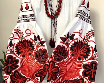 May include: White linen blouse with red and black embroidered floral patterns on the sleeves. The blouse has a V-neck and a tassel detail on the front. The text "SVIT VYSVYVANOK" is visible on the top left corner of the image.