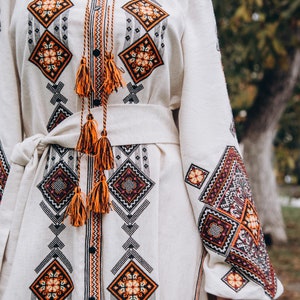 May include: A white linen dress with orange and black embroidered geometric patterns. The dress has a long sleeve and a sash tied at the waist.