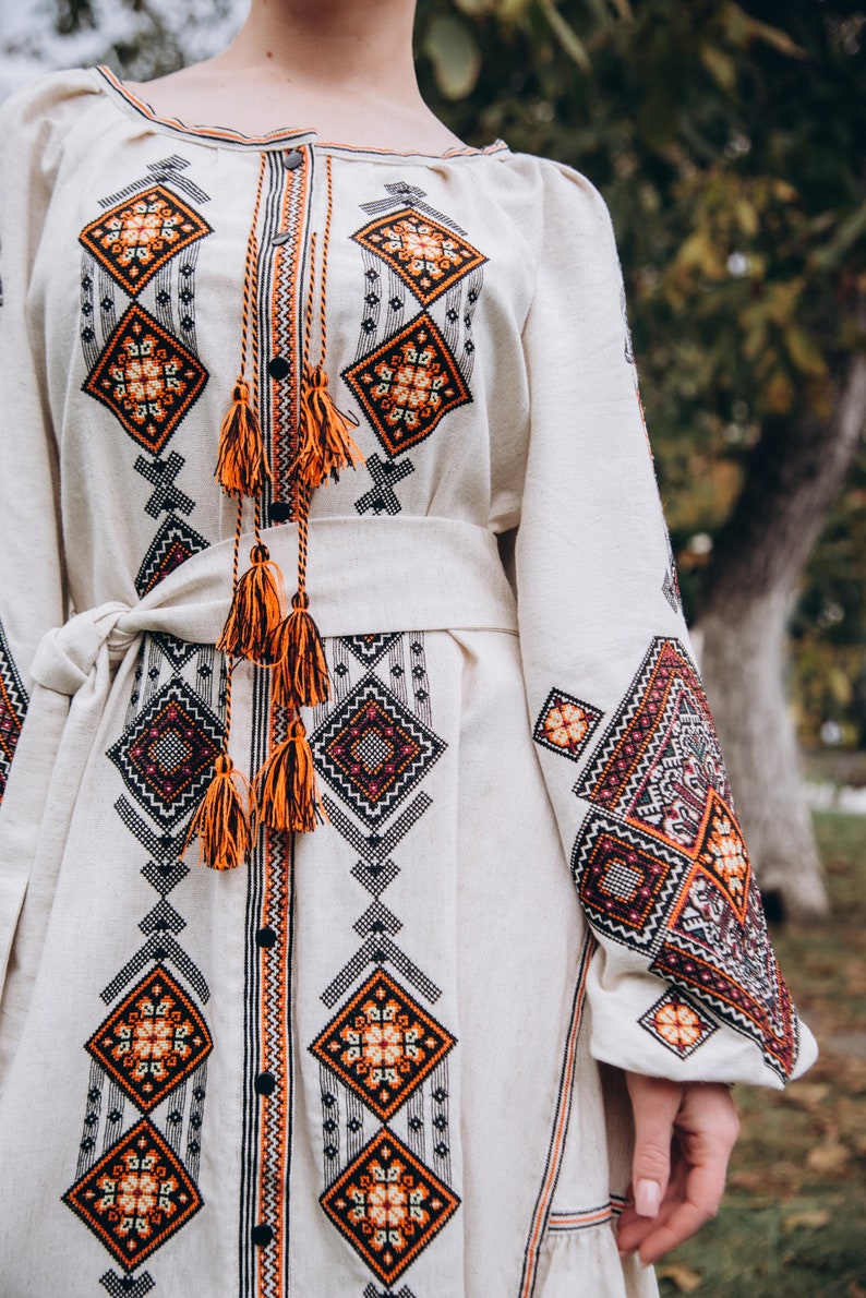 May include: A white linen dress with orange and black embroidered geometric patterns. The dress has a long sleeve and a sash tied at the waist.