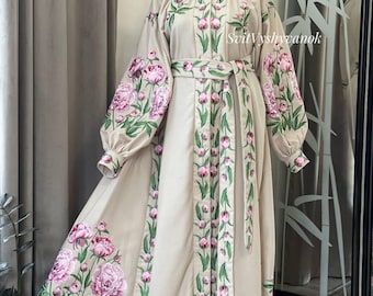 Könnte beinhalten: Beigefarbenes, langärmliges Kleid mit floralen Stickereien. Das Kleid hat rosa Pfingstrosen- und grüne Blattmuster auf den Ärmeln, dem Oberteil und dem Saum. Ein passender Gürtel betont die Taille. Der Text "SvitVyshyvanok" ist sichtbar.