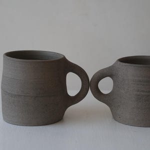 Peut inclure: Deux tasses en céramique grise avec des anses arrondies. Les tasses ont une surface texturée, légèrement rugueuse.